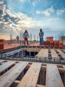 Two construction workers stand on a building site under a cloud-filled sky. Sunlight illuminates the scene.